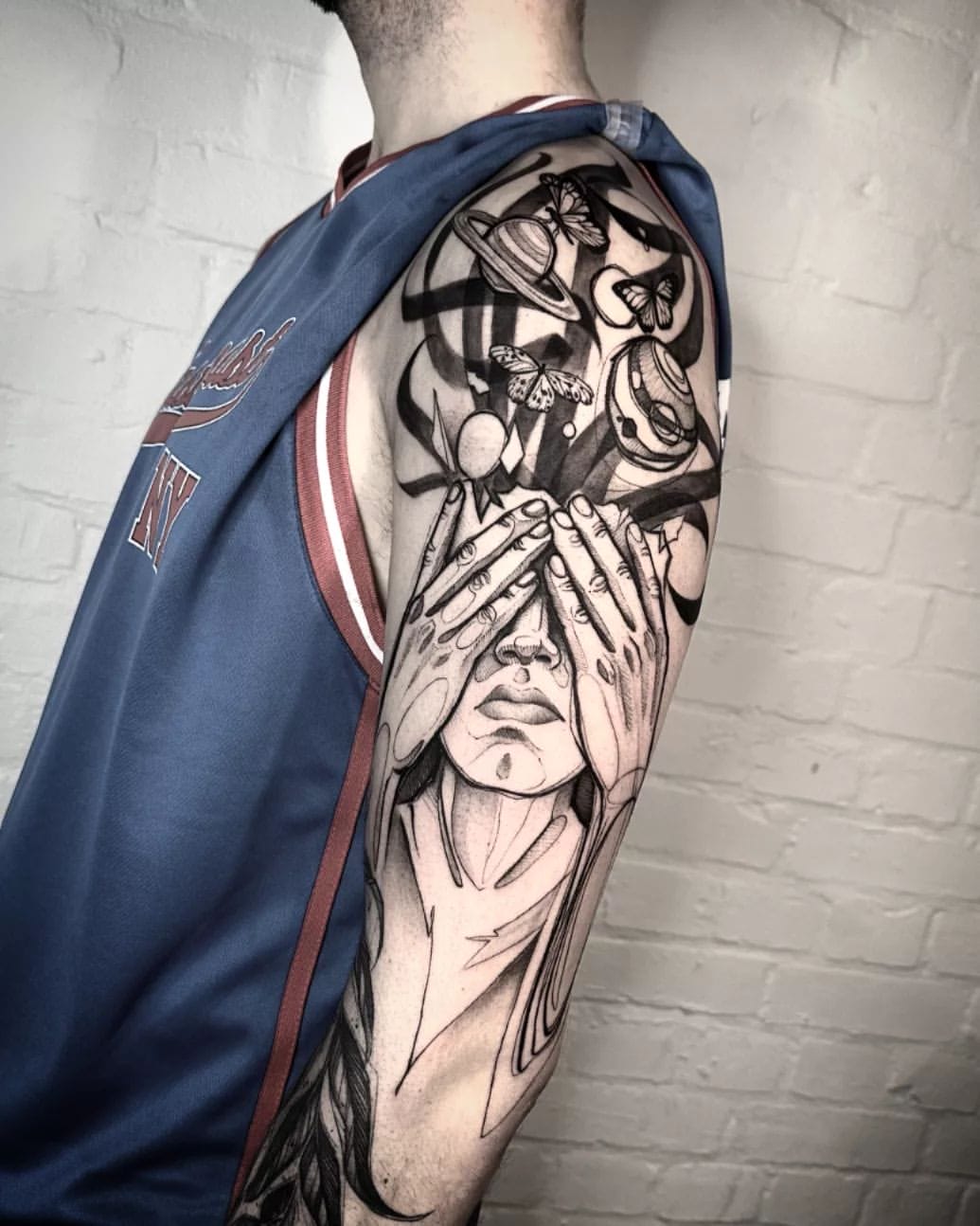 Una persona que viste una camiseta deportiva azul con detalles rojos luce un intrincado tatuaje en blanco y negro en el brazo izquierdo. El tatuaje muestra la imagen de una persona con las manos cubriéndose los ojos, rodeada de elementos celestiales y de la naturaleza, como planetas y mariposas.