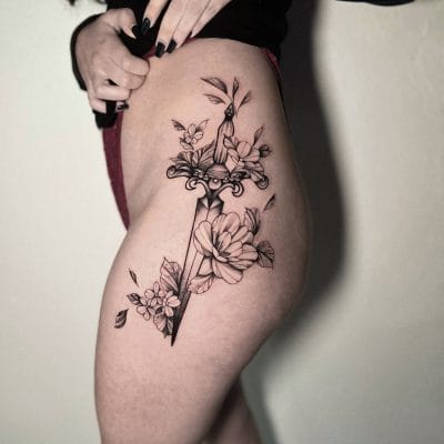 Una persona con cabello largo muestra un tatuaje detallado en tinta negra en su muslo. El diseño, elaborado en un estudio de Barcelona, presenta una daga entrelazada con flores y hojas en flor, que combina líneas y sombras intrincadas. La persona lleva una camiseta negra y pantalones cortos color granate.